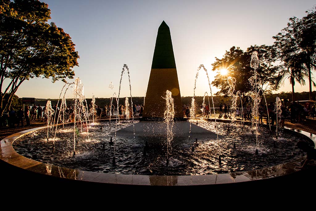 a foto mostra o marco das três fronteiras, a fonte de água e um pôr do sol.