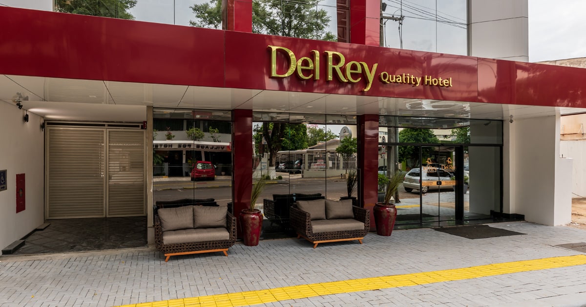 A imagem mostra a fachada do Del Rey Quality Hotel, caracterizado por um letreiro grande e dourado com o nome do estabelecimento sobre uma entrada elegante. O hotel possui uma arquitetura moderna com detalhes em vermelho e colunas robustas. À frente, há confortáveis sofás de couro posicionados para acomodar os hóspedes ou visitantes que aguardam. O local transmite uma atmosfera acolhedora e sofisticada, ideal para viajantes em busca de conforto e qualidade durante sua estadia.