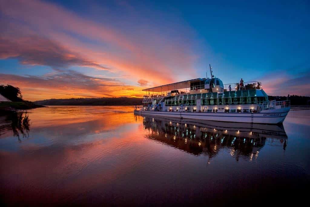 Um deslumbrante pôr do sol em Foz do Iguaçu, com o céu pintado em tons de laranja, rosa e azul profundo, refletindo nas águas calmas abaixo. O Kattamaram, uma atração turística popular na região, flutua majestosamente no lago, com luzes que começam a brilhar ao anoitecer. A cena evoca um ambiente perfeito para o Dia dos Namorados, oferecendo um cenário romântico para casais celebrarem o amor.