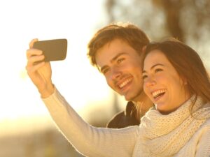 Um casal sorridente tirando uma selfie juntos, imerso na alegria e no companheirismo do Dia dos Namorados. Eles estão ao ar livre, aparentemente em um lugar iluminado pelo sol, vestidos com roupas de clima frio, sugerindo que estão em uma localidade com um ambiente fresco, tal como Foz do Iguaçu.