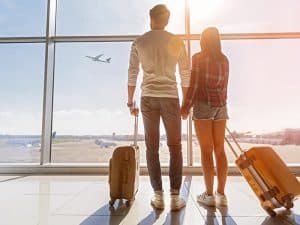 Um casal de mãos dadas está de pé em um aeroporto, olhando através de uma grande janela para um avião decolando sob um céu claro. Eles parecem estar esperando por sua própria viagem, possivelmente embarcando numa aventura romântica pelo Dia dos Namorados em Foz do Iguaçu. Ambos têm bagagem ao lado, sugerindo uma expectativa compartilhada e empolgação para o que está por vir.