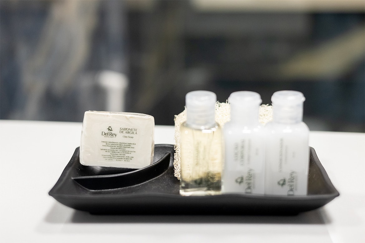 Um conjunto de amenidades de banho de cortesia da Suíte Iguassu no Del Rey Quality Hotel, apresentado com elegância. Inclui uma seleção de frascos transparentes contendo produtos de higiene pessoal como shampoo, condicionador e loção corporal, ao lado de um sabonete envolto em papel. Tudo é organizado meticulosamente em uma bandeja preta, refletindo a atenção aos detalhes e a dedicação do hotel em oferecer uma experiência excepcional aos seus hóspedes.