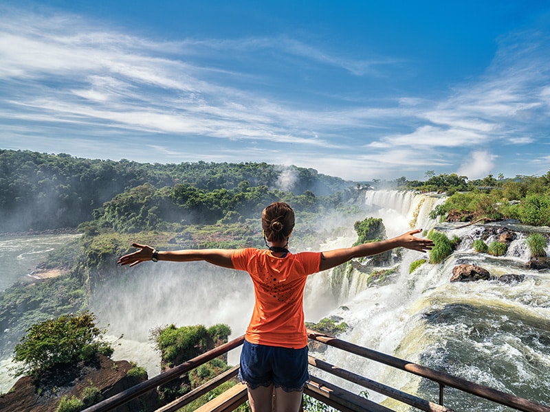 A foto mostra uma moça com os braços abertos em um pier de frente para as cataratas do Iguaçu.