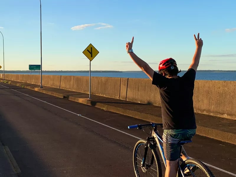 A foto mostra um ciclista com os braços levantados durante um passeio de bicicleta na Usina Hidrelétrica de Itaipu.