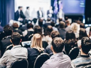 A foto mostra um congresso em andamento, com uma plateia e um palco com os palestrantes em pé.