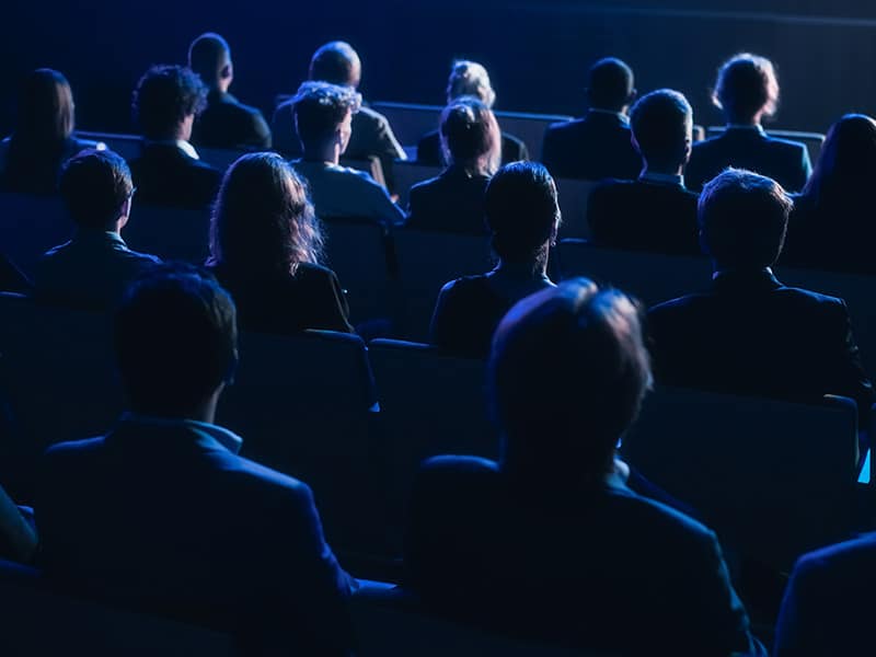 A foto mostra um auditório preenchido por pessoas, todas estão sentadas assistindo a uma palestra.