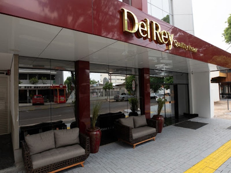 A entrada do Del Rey Quality Hotel em Foz do Iguaçu apresenta uma fachada elegante com uma marquise vinho e o nome do hotel em letras douradas. Duas colunas robustas flanqueiam a entrada, enquanto sofás de couro marrom fornecem um espaço confortável para os hóspedes relaxarem. A calçada é adornada com um tapete amarelo, convidando os visitantes a entrarem e desfrutarem da hospitalidade e conforto que o hotel oferece.