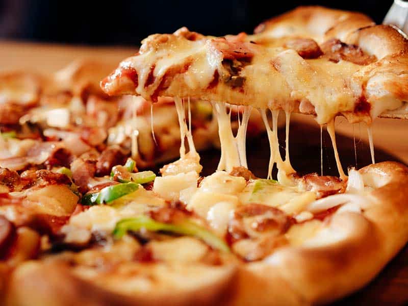 Close-up de uma suculenta fatia de pizza sendo levantada, com queijo mozzarella derretido esticando-se do restante da pizza. Coberturas incluem pedaços de bacon crocante e fatias finas de cebola, complementadas com folhas de manjericão fresco.